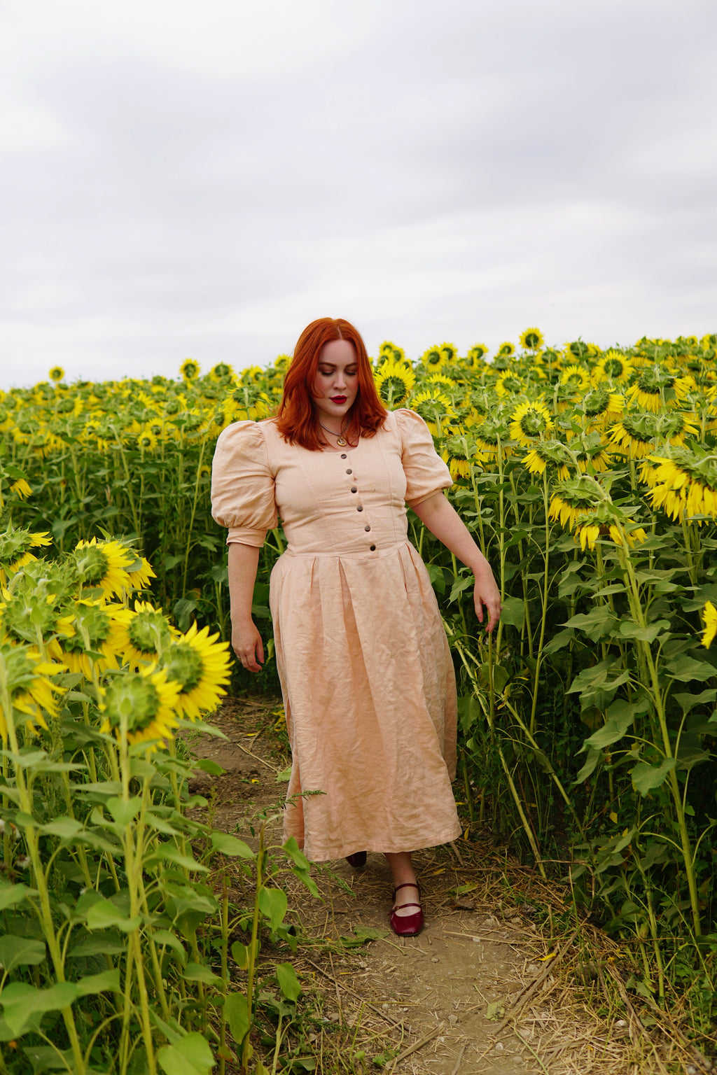 The Lobster Bisque Dress in Bisque pink organic linen, styled outdoors walking through a sunflower field