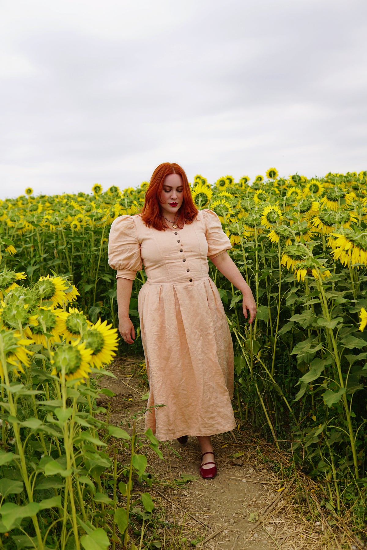 The Lobster Bisque Dress in Bisque pink organic linen, styled outdoors walking through a sunflower field