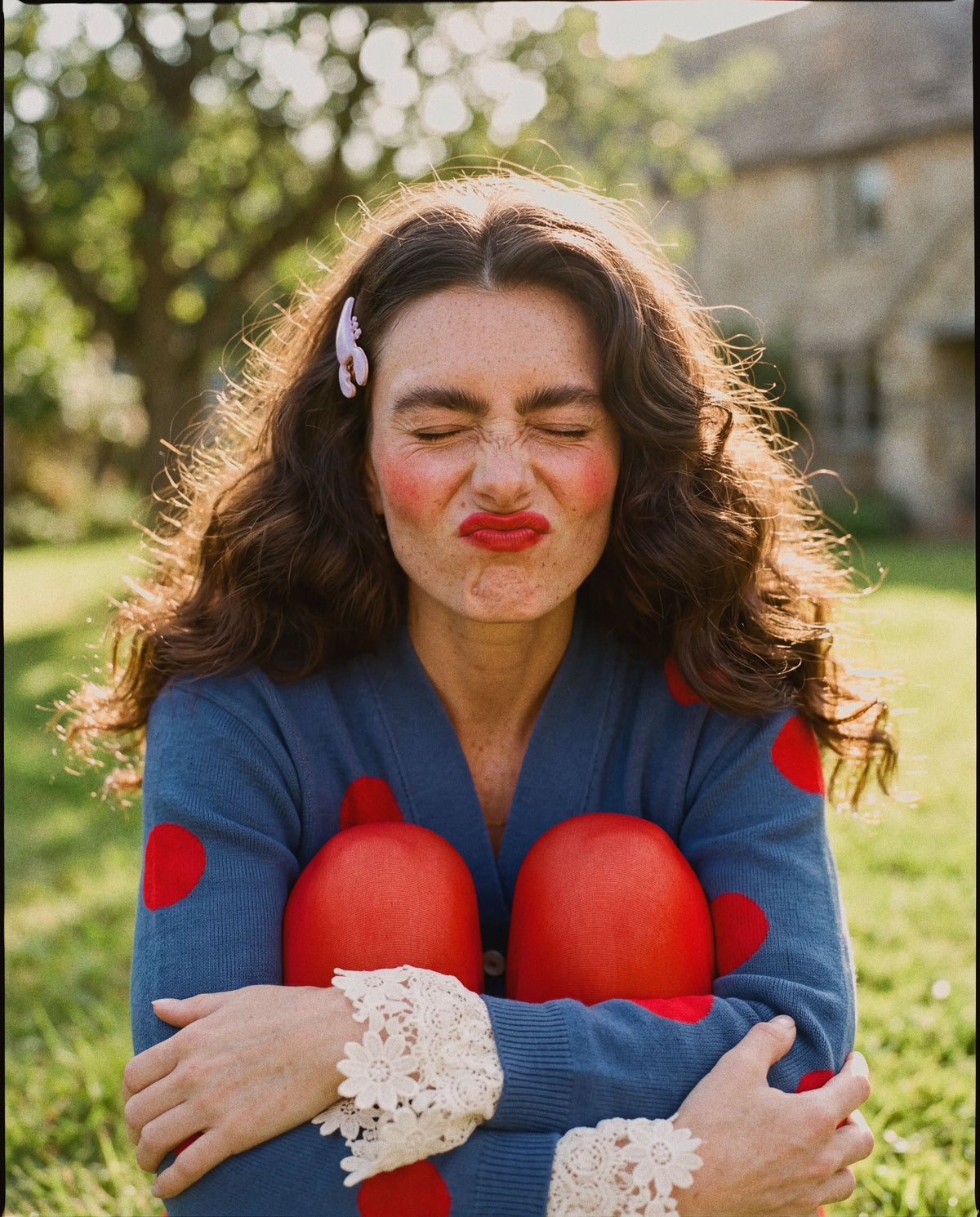 A woman outdoors in a grassy field wearing a blue cardigan with red polka dots and white lace cuffs, hugging her legs in red tights. 